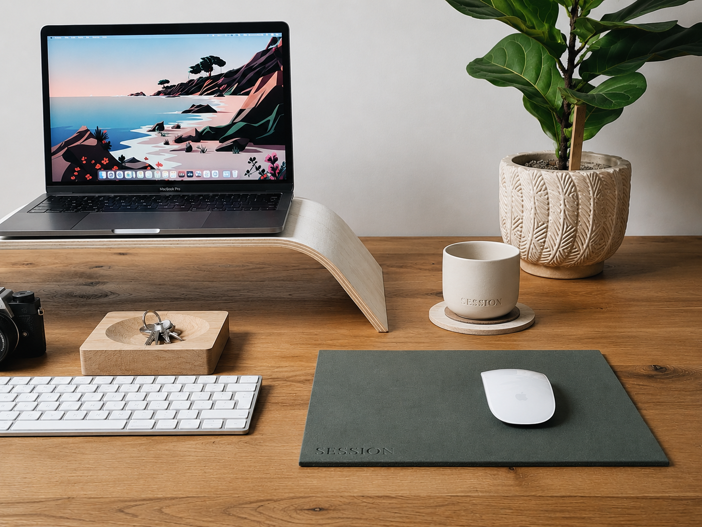 A bright minimal wooden desk with a MacBook Pro on a wooden stand, white keyboard, ceramic mug on a coaster, and fiddle-leaf plant